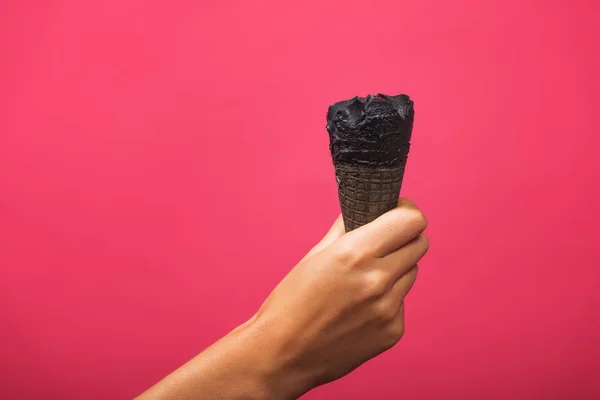 Black ice cream in a waffle cone on a pink background. Horn of coal ice cream in a female hand. Cold dessert on a colored background.