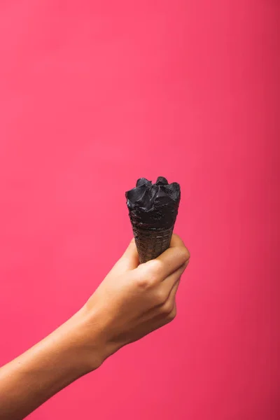 Black ice cream in a waffle cone on a pink background. Horn of coal ice cream in a female hand. Cold dessert on a colored background.