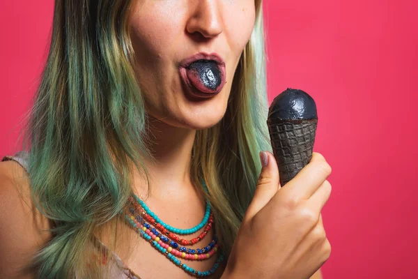 Girl with black ice cream on a pink background. Blond woman with a cold dessert. A girl with colored hair with black ice cream in a waffle cone.