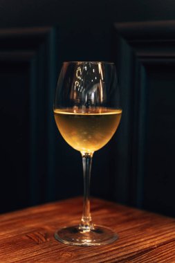 Beyaz şarap kadehleri siyah duvarda duruyor. Ahşap bir masada bir bardak içki. Cam yüzeyde parlama.