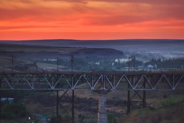 Kazakistan'da demiryolu köprüsü. Turuncu bir gün batımının arka planında yük treni. Orta Asya'da demiryolu kargo taşımacılığı. Köprü yapısı.