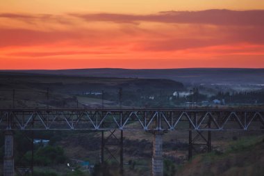 Kazakistan'da demiryolu köprüsü. Turuncu bir gün batımının arka planında yük treni. Orta Asya'da demiryolu kargo taşımacılığı. Köprü yapısı.