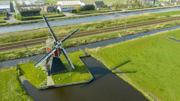 Vista aérea de un antiguo molino de viento tradicional holandés en el campo rural en los Países Bajos con un dique, canales. ferrocarril. puente y un camino . — Foto de Stock