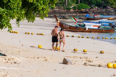 Sevgi dolu bir çift, güzel kumsalda güneşli bir günün tadını çıkarır. Kıyıda renkli renkli tekneler vardır.
