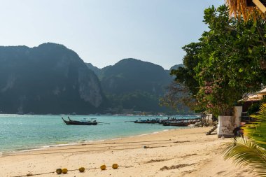 Berrak sularda yüzen, görkemli dağlarla çevrili renkli bir tekneyi tasvir eden sakin ve sakin bir sahil sahnesi.