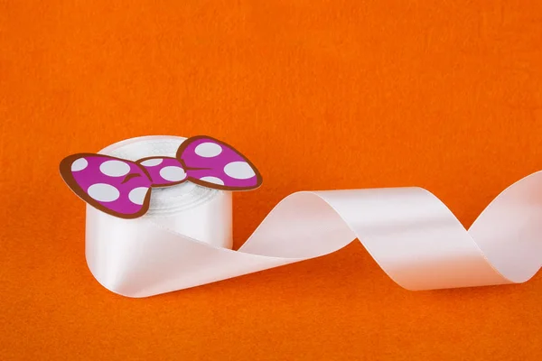 Turuncu arka planda beyaz şerit. Pembe kağıt yay. Beyaz saten kurdele. Bir rulo kaset. Dikiş atölyesi