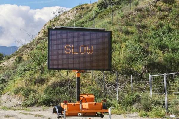 A Slow Sign on an electronic panel on the side of a freeway with a hill and clouds in the sky
