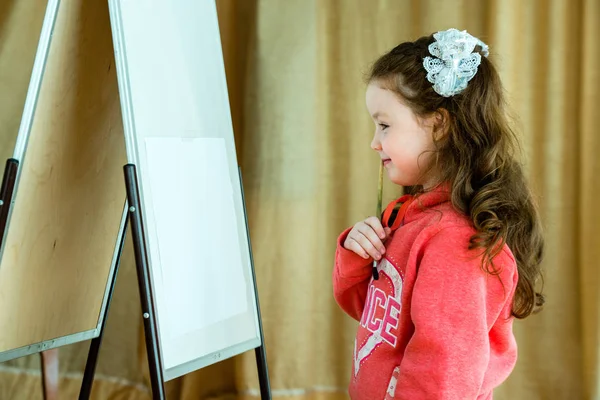 Liten flicka med en borste på staffli tänker på framtiden ritning. Vacker modell 4-5 år. — Stockfoto