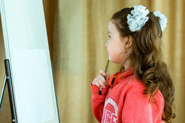 Liten flicka med en borste på staffli tänker på framtiden ritning. Vacker modell 4-5 år. — Stockfoto