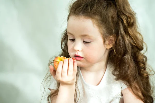 Menina bonito mordisca uma maçã com prazer. O sol brilha no rosto. Retrato emocional de uma menina bonita segurando uma maçã. Estúdio 4-5 anos . — Fotografia de Stock