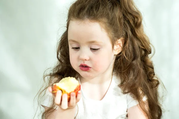 Cute little girl nibbles an apple with pleasure. Sun glare on the face. Emotional portrait of a little beautiful girl holding an apple. Studio 4-5 years. — Stock Photo, Image