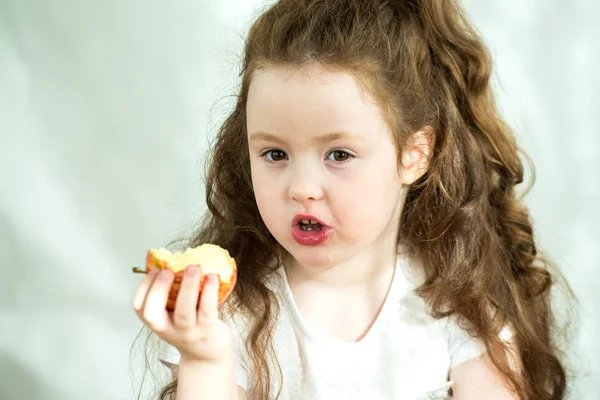 Menina bonito mordisca uma maçã com prazer. O sol brilha no rosto. Retrato emocional de uma menina bonita segurando uma maçã. Estúdio 4-5 anos . — Fotografia de Stock