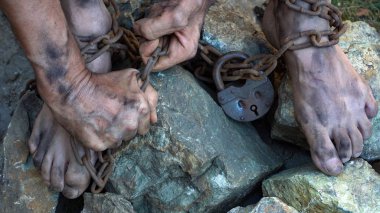 Demir zincirler içinde dolaşmış bir kölenin elleri ve ayakları. Kölelikten kurtulma girişimi. Köle emeğinin sembolü. Zincirli eller.