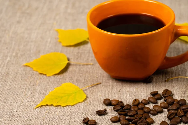 Sıcak siyah kahve bir fincan, düşmüş sarı yaprakları ve kahve çekirdekleri bir kumaş arka plan yanında. Sonbahar teması — Stok fotoğraf