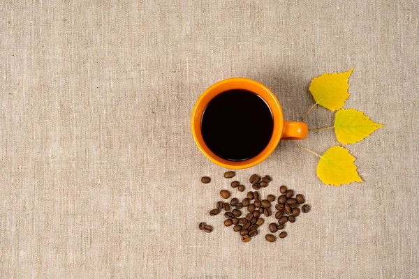 Sıcak siyah kahve bir fincan, düşmüş sarı yaprakları ve kahve çekirdekleri bir kumaş arka plan yanında. Sonbahar teması — Stok fotoğraf