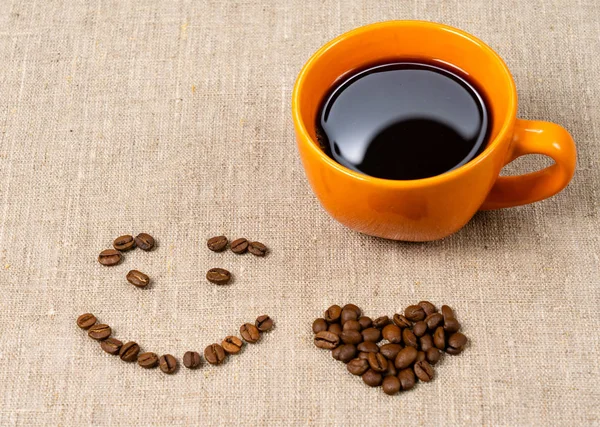 Eine Tasse heißen schwarzen Kaffee, Kaffeebohnen in Form eines Lächelns und ein Herz auf Stoffhintergrund. — Stockfoto