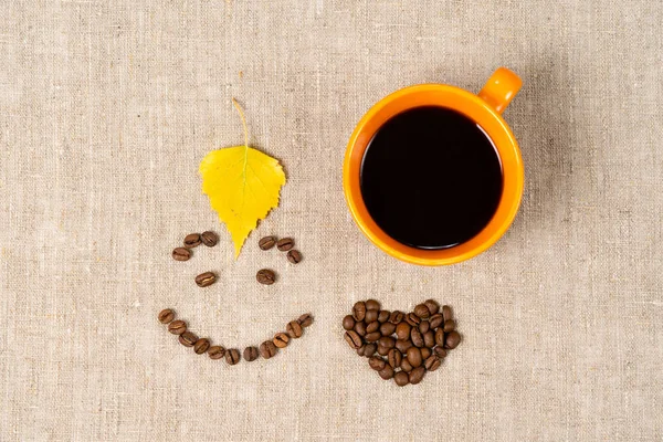 Sıcak siyah kahve bir kupa, bir gülümseme şeklinde kahve çekirdekleri ve kumaş arka plan üzerinde bir kalp. Kuru düşmüş sarı yapraklar, sonbahar teması — Stok fotoğraf