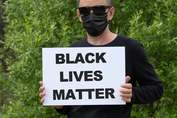 Protests broke out across the United States over the death of George Floyd. A man in a medical black mask and black glasses holds a white banner with the text BLACK LIVES MATTER.