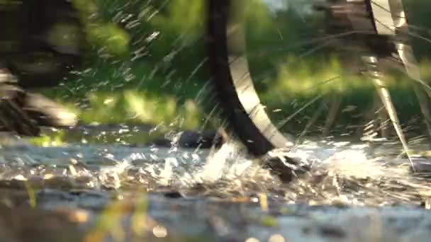 Water splashes from under the wheels.Cross the small mountain river on a mountain bike. The bike rides on the water. Shallow river in the rays of the setting sun. Slow motion, close-up, low angle, a — Stock Video