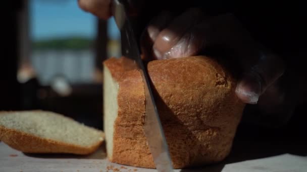 Vágj egy szelet kenyeret. Eldobható kesztyűben lévő kezek kést vágtak kenyérrel. Friss kenyér az asztalon, közel az ablakhoz, a reggeli napsütésben. Az egészséges táplálkozás és a hagyományos sütés fogalma.
