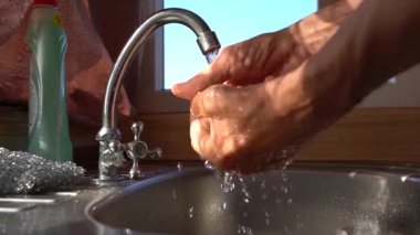 Musluğun altında ıslak eller var. Bir adam bronzlaşmış ellerini krom bir musluğun altında yıkıyor. Mutfağın penceresinde, güneş ışığıyla aydınlatılıyor.
