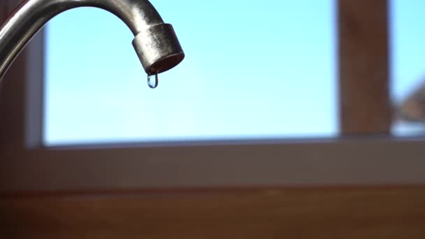 Tirar água de uma torneira ao lado de uma janela na cozinha. Gotas de água pingam rapidamente no abastecimento de água. Luz do dia. Céu azul fora da janela — Vídeo de Stock