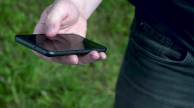Güneşli bir yaz gününde, yeşil çimenlerin arka planında bir adamın eli siyah bir akıllı telefonu tutar. Yakın plan..