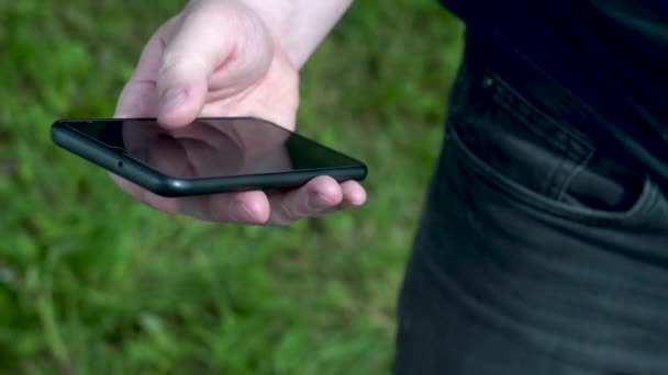 A mans hand holds a black smartphone on a background of green grass on a sunny summer day. Close-up. — Stock Video