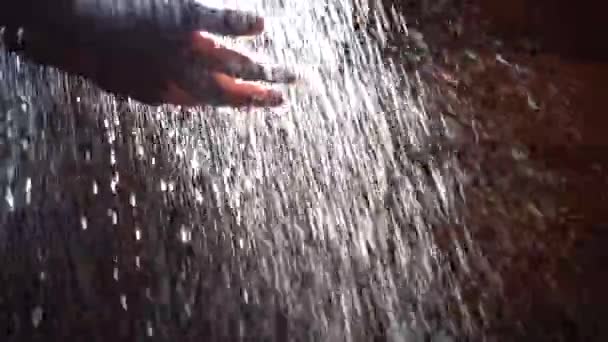 Mans hand onder de waterstralen. Achtergrondverlichting. Zonlicht. Donkere achtergrond. Jets zuiver water stromen naar beneden uit de hand van een man. Close-up — Stockvideo