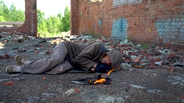 Un senzatetto dorme per strada cercando di stare al caldo vicino al fuoco. Il vagabondo giace a terra con vecchi vestiti. Il fumo di un fuoco morente punge gli occhi di un senzatetto... primo piano. Luce del giorno. — Video Stock