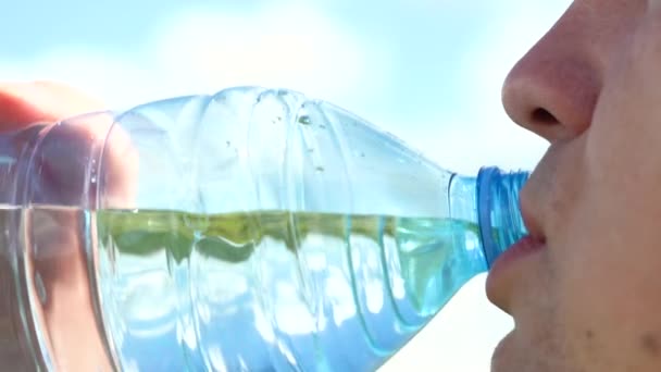 Dorst lessen. Drink schoon water uit een fles. Een jonge blanke man drinkt water uit een doorzichtige plastic fles tegen een blauwe lucht met witte wolken. Close-up — Stockvideo