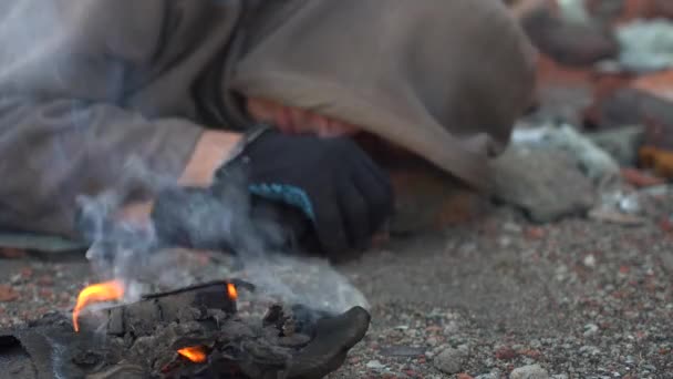 O vagabundo deita-se no chão com roupas velhas. O fumo de um incêndio moribundo pica os olhos de um sem-abrigo. Close-up. — Vídeo de Stock