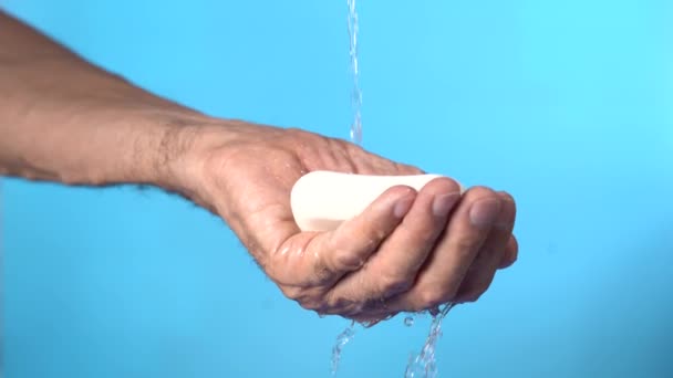 Zum Händewaschen. Männliche Hand, mit einem Stück weißer Seife unter einem Wasserstrom auf blauem Hintergrund — Stockvideo