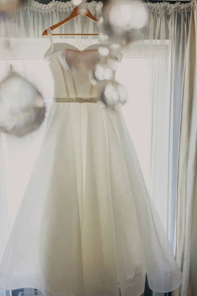 Wedding dress. White wedding dress with a full skirt on a hanger in the room of the bride with white curtains. Wedding attributes. No people.