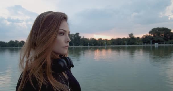 Gün Batımında Nehir Kıyısında Duran Kablosuz Kulaklıklı Güzel Genç Kadın — Stok video