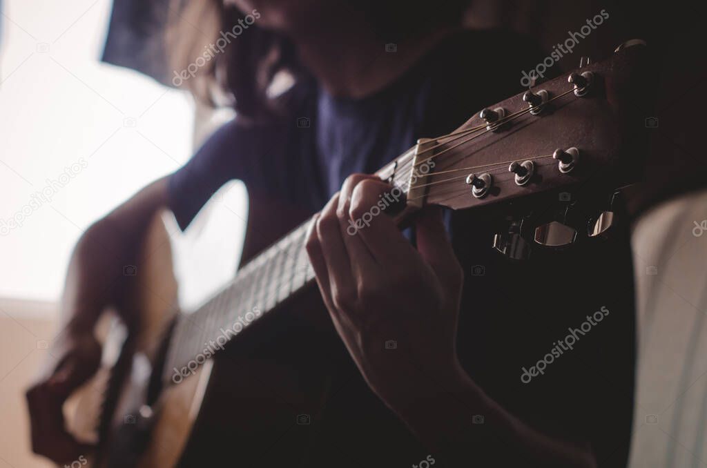 The girl plays the guitar in the room.Dim light from the window