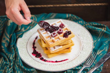Elleri kaşıkla waffle yığınına reçel döküyor. Tabakta ahududu ve pudra şekeri.