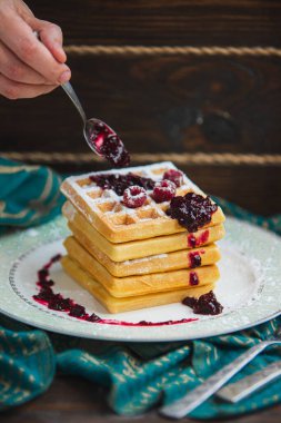Elleri kaşıkla waffle yığınına reçel döküyor. Tabakta ahududu ve pudra şekeri.