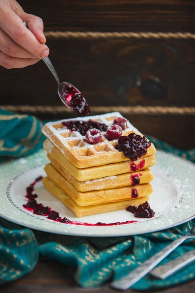 Elleri kaşıkla waffle yığınına reçel döküyor. Tabakta ahududu ve pudra şekeri.