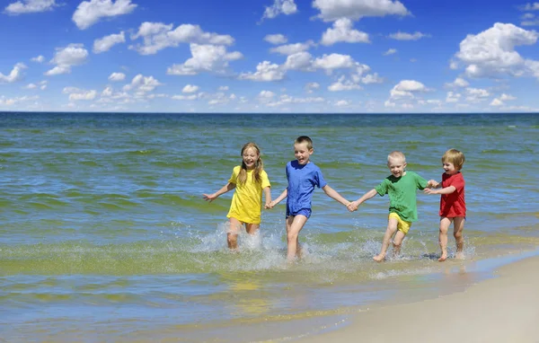 两个女孩和两个男孩在五颜六色的 T恤衫上奔跑在沙滩上 蓝天白云在后台 — 图库照片