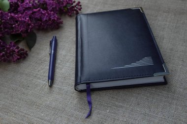 Masanın üzerindeki mavi defter ve kalemle keten bir masa örtüsü ve mor bir demet leylak.