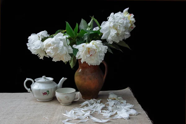 a bouquet of white peonies with green leaves in a clay jug next to a kettle and a bowl on a linen tablecloth and fallen petals on a black background