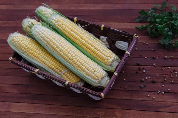 Ears of yellow corn with green leaves in a rectangular wicker basket on a dark wooden background, a table, a bunch of parsley and spilled hot pepper, peas top view