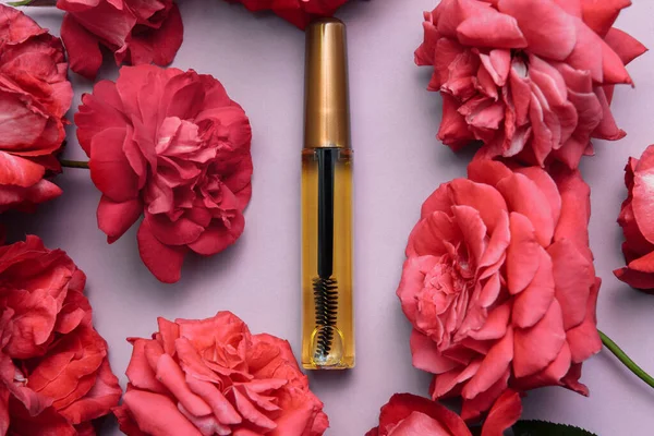 A bottle of eyelash and eyebrow oil with a black brush and a gold lid on a pink background with red rose flowers flat cosposition, the concept of grooming the eyebrows and eyelashes, restoration and g