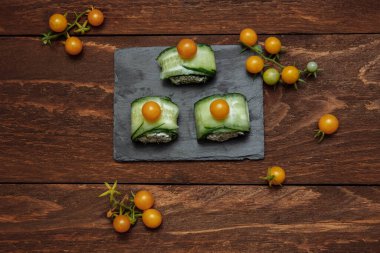 Yeşil salatalık ve peynirli sebzeli dürüm aperatifler sarı kiraz domatesli, ardoza takılmış kanepeler gri tahtada ve kahverengi ahşap arka planda servis yemekleri, yemek pişirme konsepti.