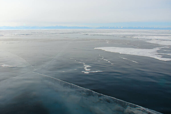 Толстый темно-синий лед пробился сквозь; замерзшие рябь темно-голубой прозрачной воды; горное озеро зимой; далекий скалистый берег утром морозный туман
