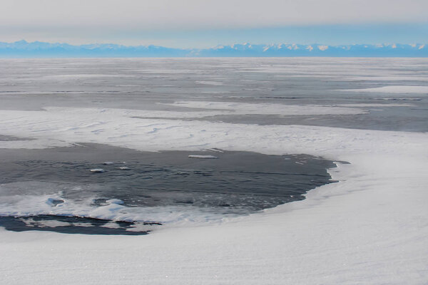 толстый темно-синий лед; замерзшие темно-голубые прозрачные воды; горное озеро зимой; далекий скалистый берег утром морозный туман
