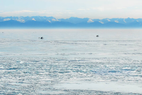 зимний пейзаж с замерзшим озером и синей горной цепью вдалеке; два судна на воздушной подушке посреди ледяного поля

