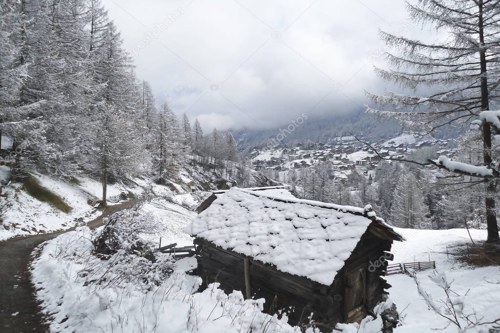 alpine landscape with an old traditional wooden shed nearby dirt road running along the mountain slope; low fluffy cloud over a distant highland village, winter in Swiss Alps