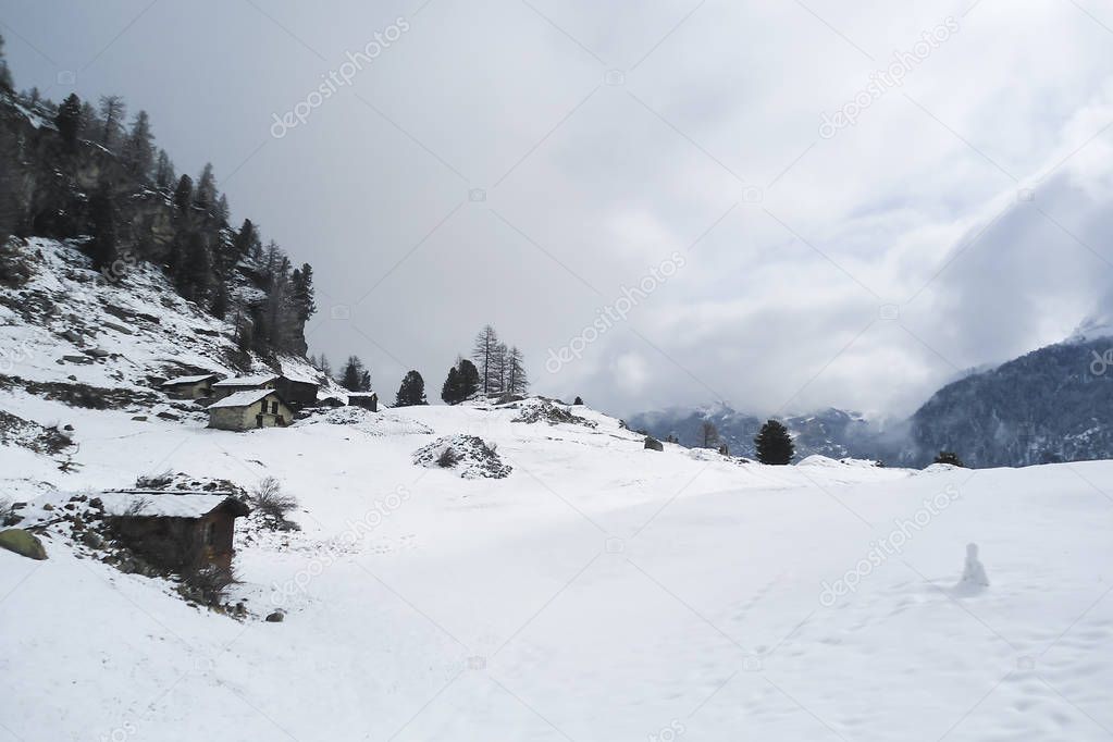 mountain landscape with old sheds and a little snowman in the middle of field in outskirts of an alpine village, winter in Swiss Alps